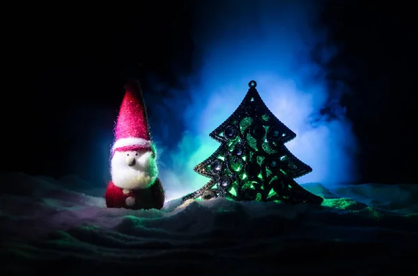 हैप्पी सांता क्लॉस गुड़िया क्रिसमस के समय पेड़ और बर्फ के साथ। रंगीन बोकेह पृष्ठभूमि। सांता क्लॉज और मेरी क्रिसमस मॉडल फिगर खिलौना डार्क टोन धुंधली पृष्ठभूमि पर — स्टॉक फ़ोटो, इमेज