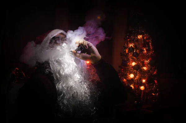 Santa Clause vaping electronic cigarette dressed as traditional Santa on a dark toned background with vape clouds. Selective focus