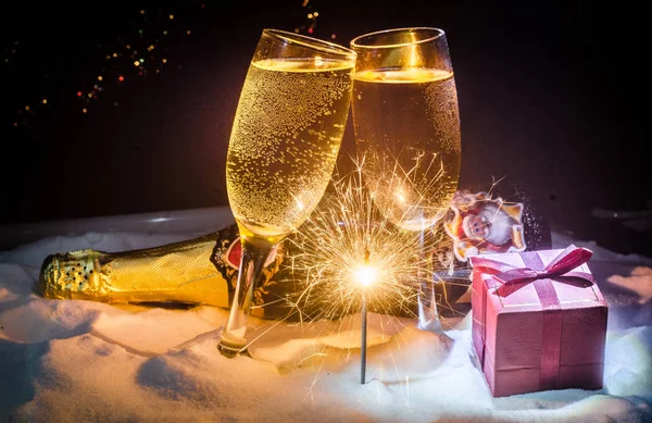 New Year Eve celebration background with pair of flutes and bottle of champagne with Christmas attributes (or elements) on snowy dark toned foggy background. Selective focus. Useful as greeting card