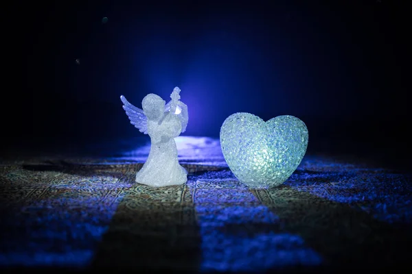 Статуэтка маленького ангела с сердцем и обручальными кольцами на темно-синем фоне . — стоковое фото