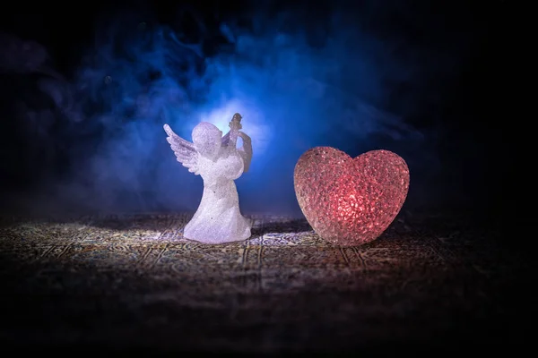 Statuette de petit ange et coeur et anneaux de mariage sur fond brouillard bleu foncé . — Photo