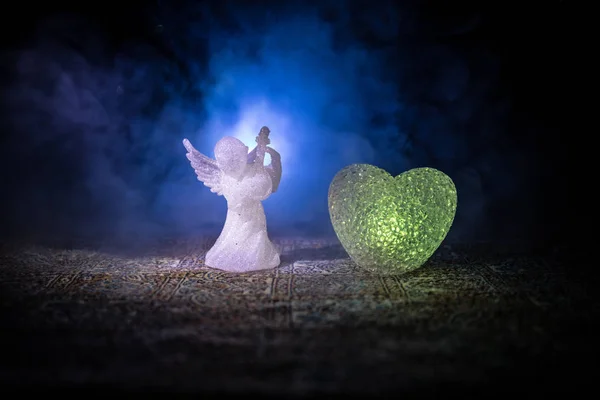 Estatueta de pequeno anjo e coração e anéis de casamento sobre fundo nevoeiro azul escuro . — Fotografia de Stock