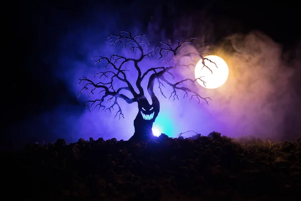 Silhouette of scary Halloween tree with horror face on dark foggy toned background with moon on back side. Scary horror tree with zombie and demon faces.