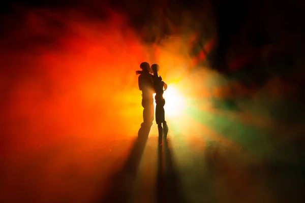 Love Valentine`s Day concept.Sillhouette of sweet young couple in love standing in the field and hugging on dark toned foggy background. Decoration with doll figures on table shot. — Stock Photo, Image