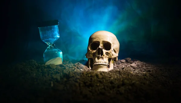 Totenkopf und Vintage-Sanduhr auf dunkel getöntem Nebelhintergrund im Lichtkegel. Horror-Konzept. — Stockfoto