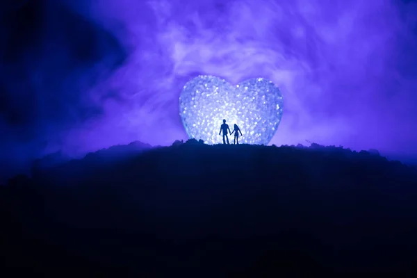 Silhouet van gelukkige paar staande achter grote gevormde hartsymbool op berg 's nachts. Groot hart zoals maan gloeien op mistige hemel. Aftelkalender voor Valentijnsdag decor foto — Stockfoto