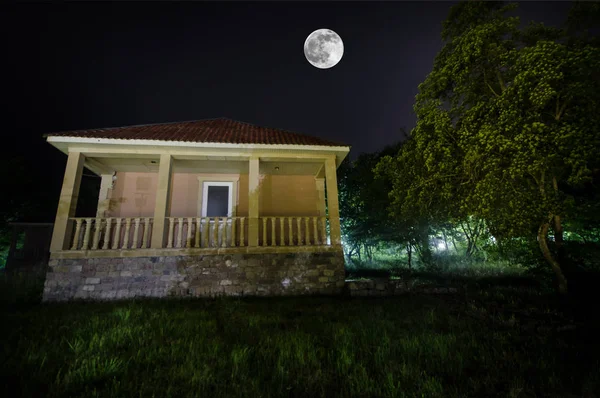 Horská noční krajina budovy v lese v mlhavé noci s měsícem. Zelená louka, velké stromy a opuštěného domu v noci. Noční scéna — Stock fotografie