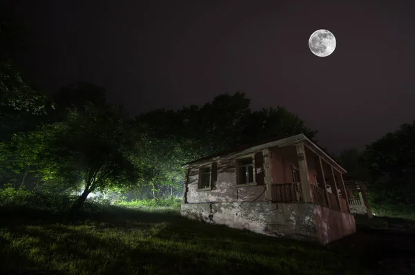Горный ночной пейзаж постройки в лесу в туманную ночь с луной. Зеленый луг, большие деревья и заброшенный дом ночью. Ночная сцена — стоковое фото