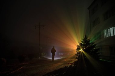 Gece yoğun sis içinde şehir. Mistik manzara ürkütücü adam gerçeküstü ışıklarla. Yapay ışıkta gece siste yürüyen adamın siluet. Arka aydınlatma karışık güzel.
