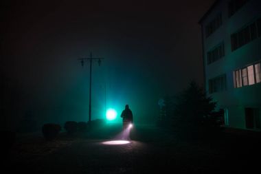 Gece yoğun sis içinde şehir. Mistik manzara ürkütücü adam gerçeküstü ışıklarla. Yapay ışıkta gece siste yürüyen adamın siluet. Arka aydınlatma karışık güzel.