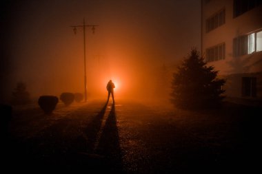 Gece yoğun sis içinde şehir. Mistik manzara ürkütücü adam gerçeküstü ışıklarla. Yapay ışıkta gece siste yürüyen adamın siluet. Arka aydınlatma karışık güzel.