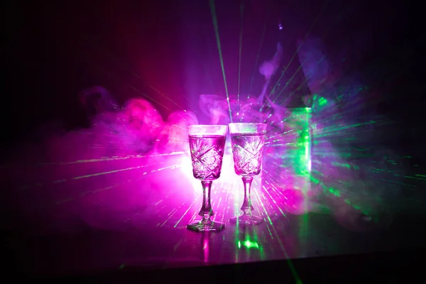 Dois copos de Vodka com garrafa em fundo estilo clube nevoeiro escuro com luzes brilhantes (Laser, Stobe) Multi colorido. Clube bebidas tema decoração. Espaço vazio. — Fotografia de Stock