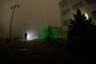 Gece yoğun sis içinde şehir. Mistik manzara ürkütücü adam gerçeküstü ışıklarla. Yapay ışıkta gece siste yürüyen adamın siluet. Arka aydınlatma karışık güzel.