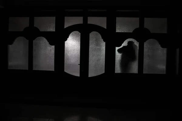 Silhouette di una figura ombra sconosciuta su una porta attraverso una porta a vetri chiusa. La silhouette di un umano davanti a una finestra di notte. Scena spaventosa concetto di Halloween di silhouette sfocata — Foto Stock