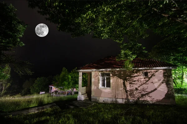 Montanha noite paisagem de construção na floresta em noite nebulosa com a lua. Prado verde, árvores grandes e casa abandonada à noite. Cena noturna — Fotografia de Stock