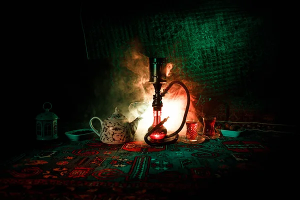Кальян с горячими углями на чаше с шишами делает облака пара в арабском интерьере. Восточный орнамент на ковре восточной чайной церемонии. Стильный восточный шиш в темноте с подсветкой . — стоковое фото