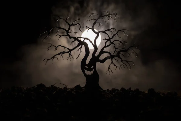 Silhouette of scary Halloween tree with horror face on dark foggy toned background with moon on back side. Scary horror tree with zombie and demon faces.