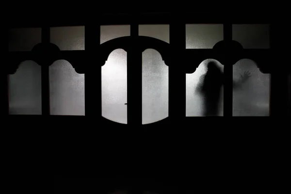 Silueta de una figura de sombra desconocida en una puerta a través de una puerta de cristal cerrada. La silueta de un humano frente a una ventana por la noche. Escena de miedo concepto de Halloween de silueta borrosa —  Fotos de Stock