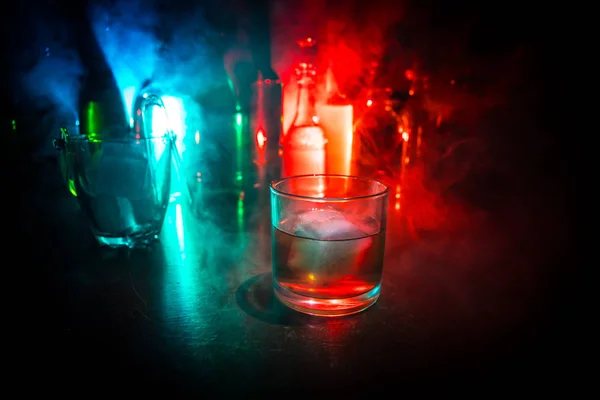 Bicchiere di whisky su barra di legno primo piano con bottiglie vista offuscata su sfondo scuro con luce e fumo. Bicchiere singolo di whisky su ghiaccio con una superficie di legno riflettente — Foto Stock