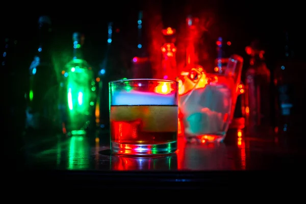 Verre de whisky sur bar en bois gros plan avec des bouteilles vue floue sur fond sombre avec lumière et fumée. Un seul verre de whisky sur glace avec une surface réfléchissante en bois — Photo