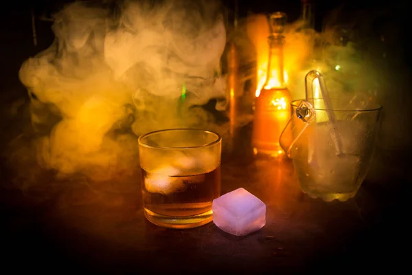 Sklenice whisky na dřevěný bar detailní s lahví rozmazaný pohled na tmavém pozadí se světlem a kouř. Jednu sklenici whisky na ledě s reflexním povrchem, dřevěné — Stock fotografie