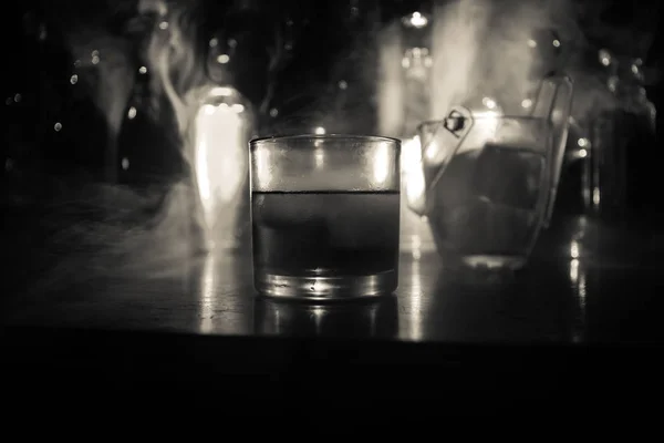 木製のバー クローズ アップをボトルにウイスキーのガラスは 光と煙で暗い背景にビューをぼやけています 反射型木製の表面が付いている氷でウイスキーの つのガラス 選択と集中 サービスを提供する準備ができて — ストック写真