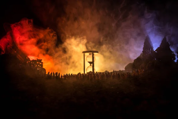 Silhouette einer großen Menschenmenge im Wald bei Nacht vor einer großen Sanduhr mit getönten Lichtstrahlen auf nebligem Hintergrund. Zeitkonzept. — Stockfoto