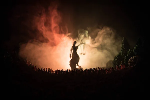 Silhouette de dame géante floue statue de justice avec épée et balance debout derrière la foule la nuit avec fond de feu brumeux . — Photo