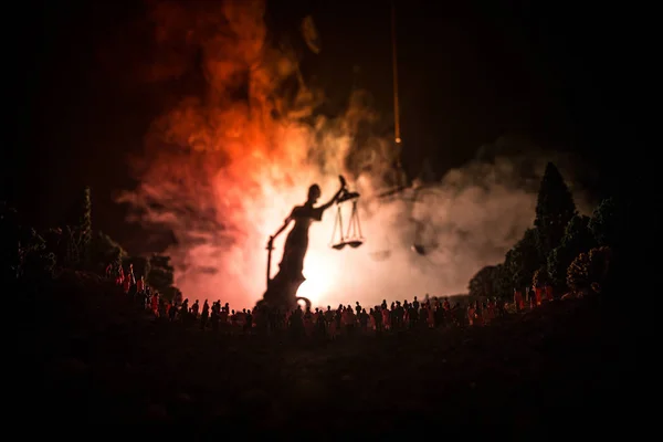 Silueta rozmazané obří dáma socha spravedlnosti s mečem a měřítka stojící za dav v noci s pozadím mlhavé oheň. — Stock fotografie