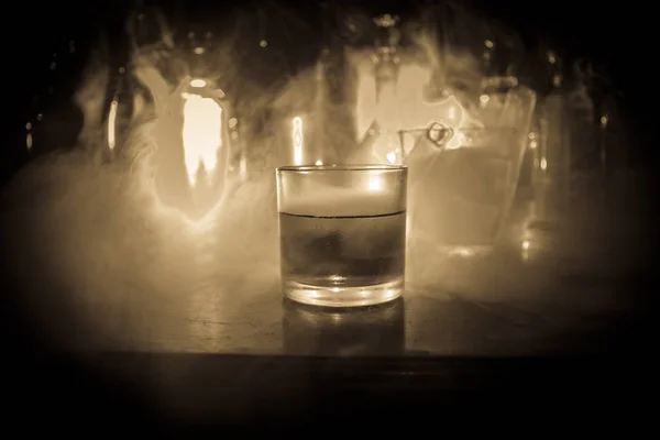 玻璃在木酒吧特写与瓶子模糊的看法在黑暗背景与光和烟雾 冰上的单杯威士忌 有反光的木质表面 选择性对焦 准备服务 — 图库照片
