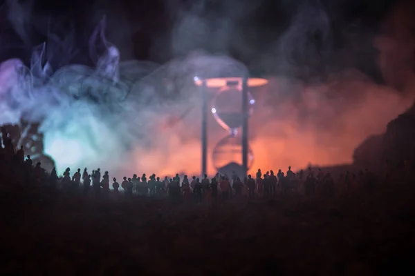 Silueta de una gran multitud de personas en el bosque por la noche de pie contra un reloj de arena grande con rayos de luz tonificados sobre fondo de niebla. Concepto de tiempo . — Foto de Stock