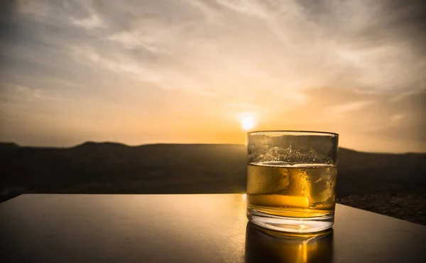 Un bicchiere di whisky con ghiaccio su sfondo tramonto o un bicchierino di whisky al tramonto drammatico cielo su sfondo paesaggio montano — Foto Stock