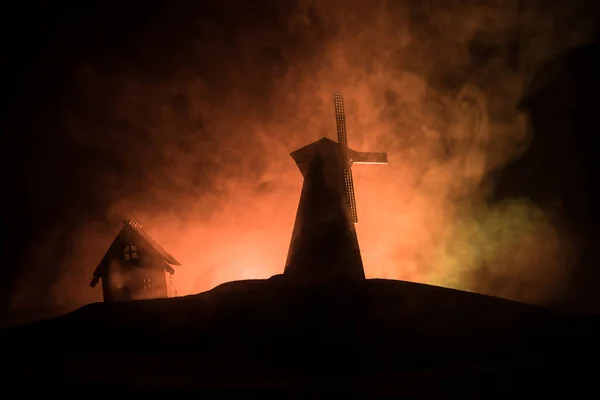 Silueta del molino de viento de pie en la colina contra el cielo nocturno. Decoración nocturna con viejo molino de viento en la colina con fondo nebuloso de horror tonificado con luz . — Foto de Stock