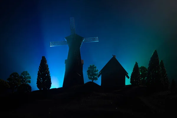 Silhueta de moinho de vento em pé na colina contra o céu noturno. Decoração noturna com moinho de vento velho na colina com horror tonificado fundo nebuloso com luz . — Fotografia de Stock