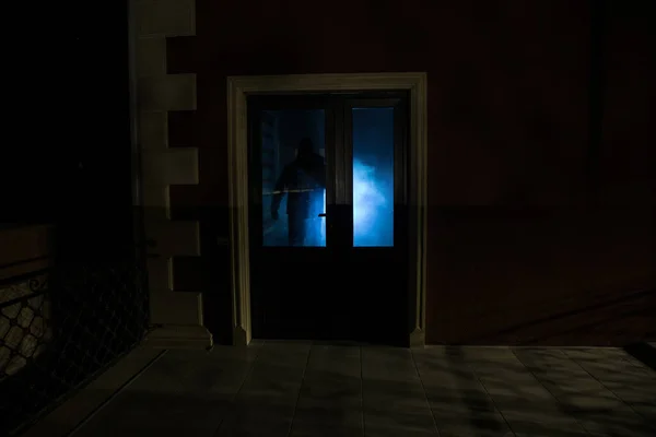 Silhouette of an unknown shadow figure on a door through a closed glass door. The silhouette of a human in front of a window at night. Scary scene halloween concept of blurred silhouette of maniac.