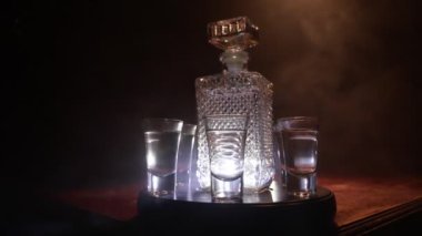 A beautiful carafe with vodka stands on a wooden table with glasses. Dramatic toned foggy background with light. Selective focus. Club drink concept.