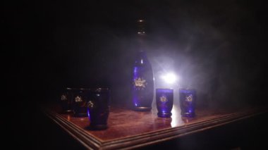 A beautiful blue carafe with vodka stands on a wooden table with glasses. Dramatic toned foggy background with light. Selective focus. Club drink concept.