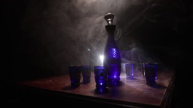A beautiful blue carafe with vodka stands on a wooden table with glasses. Dramatic toned foggy background with light. Selective focus. Club drink concept.