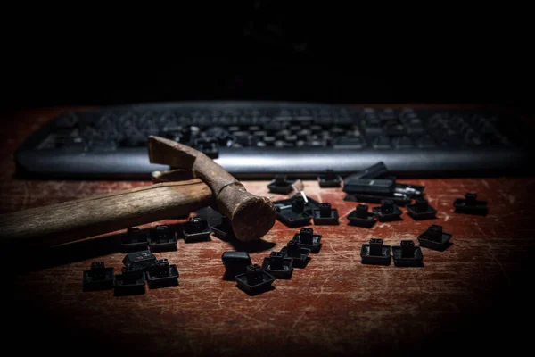 Broken keyboard with hammer on wooden table. Keyboard with no keys on dark desktop.