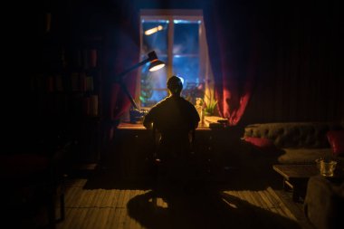 Geceleri mobilyası ve penceresi olan gerçekçi bir bebek evi oturma odası. Karanlık odada masada oturan bir adam. Seçici odak.