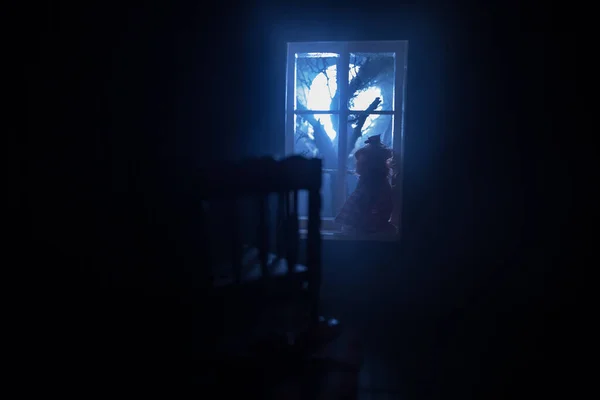 Old creepy eerie baby crib near window in dark room. Scary baby silhouette in dark. A realistic dollhouse living room with furniture and window at night. Selective focus