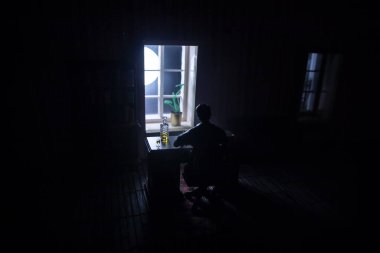 Geceleri mobilyası ve penceresi olan gerçekçi bir bebek evi oturma odası. Karanlık odada masada oturan bir adam. Küresel virüs salgını sırasında evde kalma kavramı. Seçici odak.