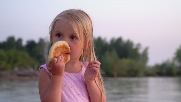 La petite fille aux cheveux blonds sur la plage. La petite fille mange rouler lors d'un déclin à la mer — Video