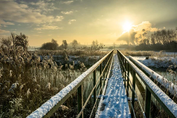Зимний пейзаж с пешеходным мостом через реку, современная архитектура в сельской местности и мрачное небо заката — стоковое фото