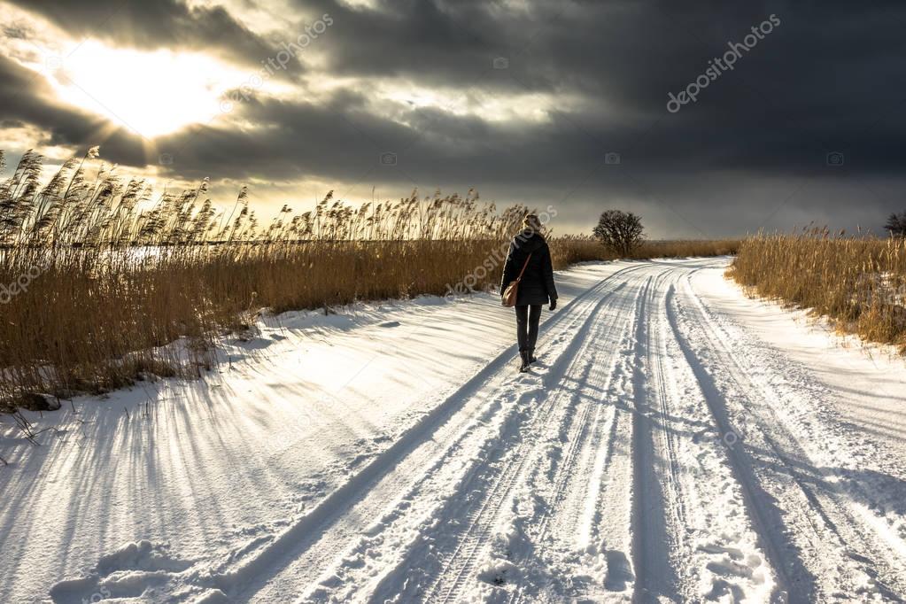 Walking woman in winter landscape, road with snow, moody sky before sunset by the lake