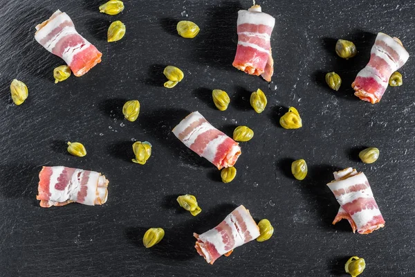Smoked bacon, rolled meat as tapas or antipasto with capers served on black slate