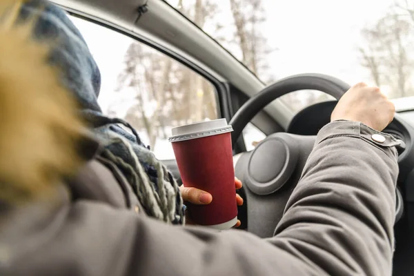 Kierowca pijący kawę w samochodzie, prowadzący i trzymający kierownicę oraz filiżankę gorącego napoju w chłodny zimowy dzień ze śniegiem za oknem — Zdjęcie stockowe