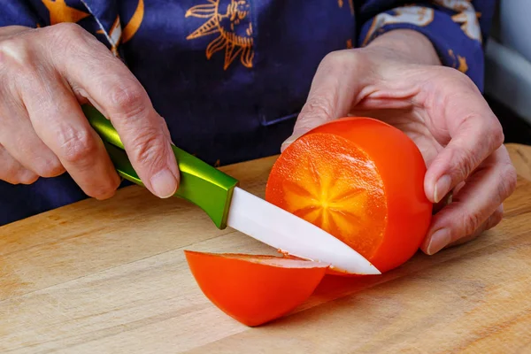 Літня жінка порізає стиглий оранжевий хуртовина керамічним ножем на дерев'яній обробній дошці. Проклята хурма в руці літньої жінки крупним планом — стокове фото