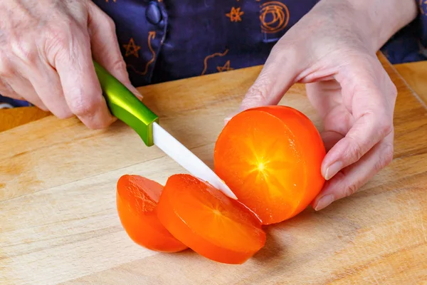 Mulher idosa corta cáqui suculento laranja brilhante por faca de cerâmica na placa de corte de madeira. Vista superior close-up — Fotografia de Stock