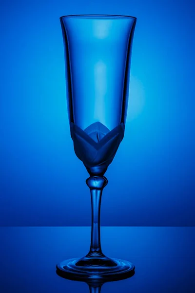 Empty transparent champagne glass on a thin high leg in dark blue backlight with reflection on a glossy surface — Stock Photo, Image